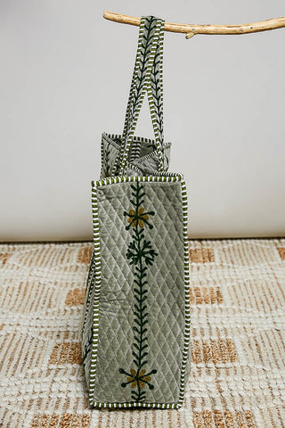 A light gray quilted tote bag with green and white striped handles and a vertical green floral embroidery, hanging from a wooden branch over a textured beige rug.