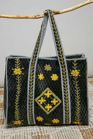 A black quilted tote bag with yellow floral embroidery and white-and-black striped handles, hanging from a natural wood branch against a light gray background.