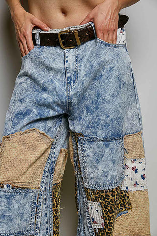 A close-up of a person wearing heavily patchwork-style blue denim jeans with burlap, leopard-print, floral, and white patch panels, plus a dark belt with a gold buckle.