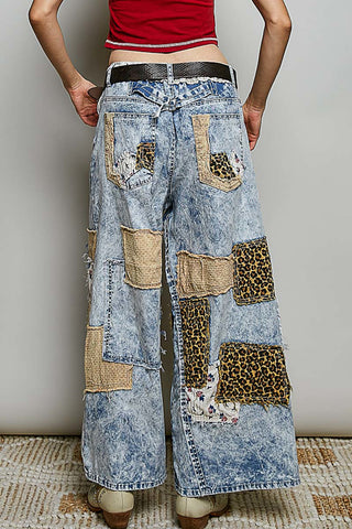 A person shown from the back wearing wide-leg patchwork denim jeans with a blue acid-wash base and multiple tan, leopard-print, and floral fabric patches, paired with a red top and brown shoes.