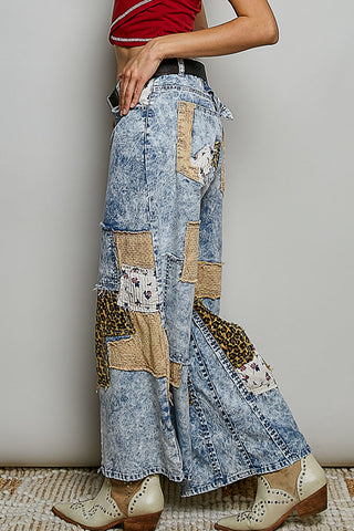 A person wearing wide-leg patchwork denim pants with light blue washed denim, tan fabric panels, leopard print patches, and floral applique, paired with cream western-style boots against a plain light gray wall.