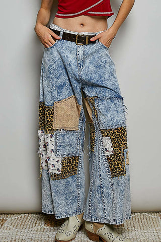 A person wearing wide-leg cropped denim pants with a blue acid-wash finish and mixed animal-print patchwork panels, paired with a brown belt and light-colored clogs, standing against a plain light gray wall.
