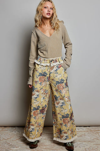 A woman modeling a beige long-sleeve knit top and wide-leg floral pants with ruffled trim, standing against a plain light gray wall.