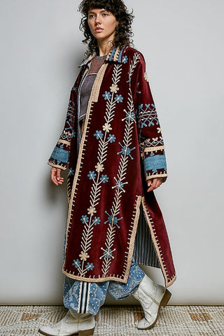 A woman models a long, bohemian-style maroon velvet coat with intricate blue, beige, and white floral embroidery, paired with patterned pants and white boots against a neutral studio background.