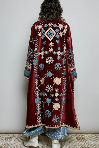 Person wearing a long, embroidered burgundy coat with floral patterns on a plain background
