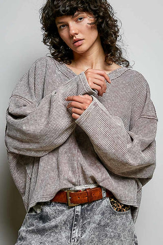 A model wearing a loose, oversized long-sleeve top with a fine black and white micro-check pattern, styled with a brown leather belt and gray acid-wash jeans against a plain light gray background.