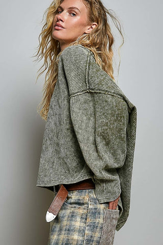 A woman wearing a cropped, oversized houndstooth jacket in olive green and beige, paired with a brown belt and plaid pants, photographed against a neutral gray studio background.