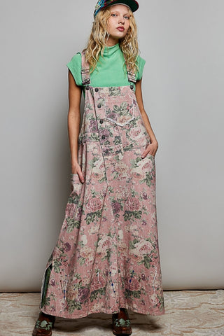 A woman modeling a long pink floral overall dress with a green t-shirt underneath, a patterned cap, and brown shoes, standing against a plain light gray wall.