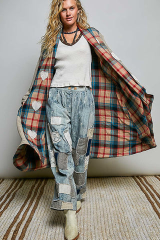 A woman models a long plaid coat with heart-shaped patch details on the front and back. She is wearing a white ribbed top, light blue patchwork denim pants, and beige boots, standing on a striped rug against a neutral gray wall.