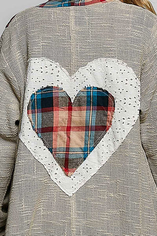 Close-up of the back of a textured beige knit sweater featuring a large white heart applique with a central plaid heart patch in red, blue, and tan.