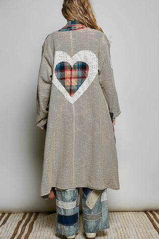 A person shown from the back wearing a long taupe-gray patchwork cardigan with a large white heart applique and a red plaid heart design in the center, paired with blue denim patchwork pants against a neutral studio background.