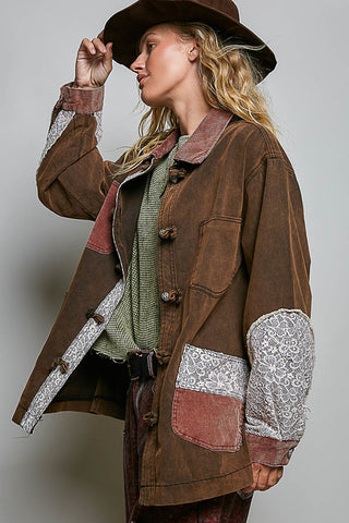 A woman wearing a brown patchwork jacket with lace-trimmed elbow patches, a brown hat, and a green knit sweater, posed against a plain light gray studio background.