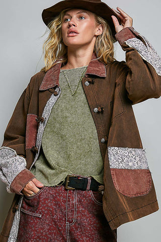 A woman wearing a brown patchwork jacket with lace-trimmed cuffs and pockets, a green patterned top, and a brown hat, photographed against a plain light gray studio background.