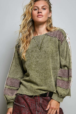 A woman wearing a long-sleeve textured knit top in olive green with patchwork details in mauve and beige on the shoulders and sleeves, paired with patterned red pants, against a plain light gray studio background.