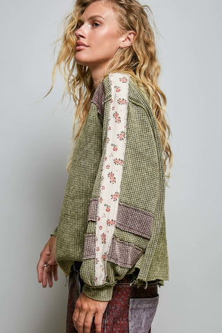 A woman wearing a patchwork-style green and white patterned jacket with floral trim and patch pockets, paired with patterned pants, photographed against a plain light gray studio background.