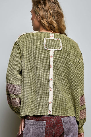 A woman viewed from the back wearing a cropped, patchwork-style green top with a textured grid pattern, a vertical cream floral panel, and patchwork fabric details on the sleeves and lower back.