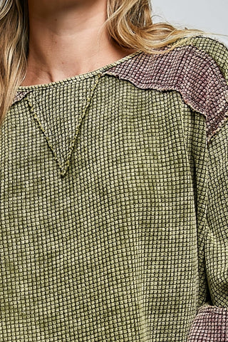 Close-up of a textured olive green top with a pinkish-purple waffle-knit yoke and neckline detail, worn by a model.
