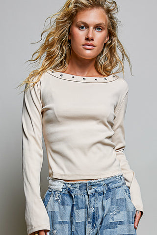 A woman wearing a cream-colored long-sleeve crop top with silver stud detailing around the wide neckline, paired with blue patchwork jeans, against a solid gray studio background.