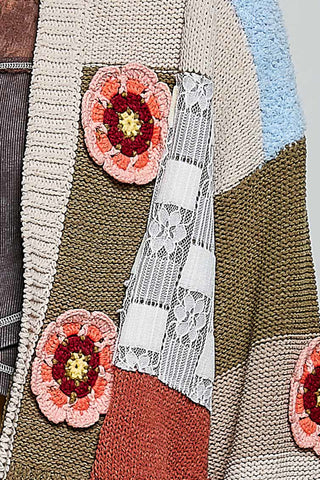 Close-up of a patchwork knit cardigan featuring textured knit panels in beige, brown, light blue, and rust orange, with white floral lace inserts and pink, red, and yellow crocheted flower appliqués.