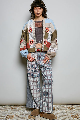 A model stands against a light gray wall wearing a patchwork knit cardigan with floral motifs, matching wide-leg patterned pants, and brown suede boots.