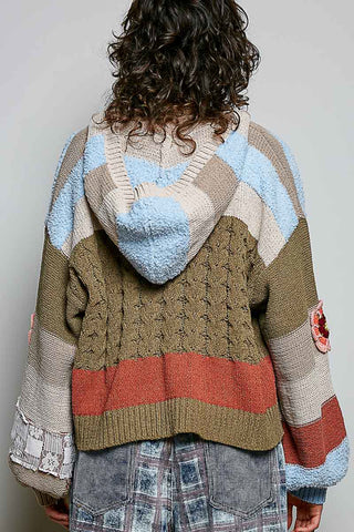 A person shown from the back wearing a chunky, multi-colored patchwork knit hoodie with a hood. The hoodie features stripes of light blue, beige, brown, rust red, and gray, with textured knit and cable knit patterns.