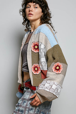 A woman wearing a patchwork knit cardigan with crochet flower appliqués, styled with a dark crop top and plaid jeans against a plain gray studio background.