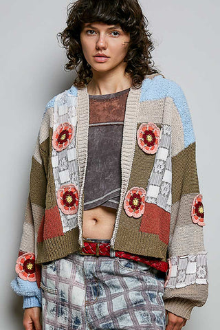 A model wearing a chunky knit patchwork cardigan with colorful floral appliqués, layered over a cropped brown top and paired with plaid pants, against a neutral gray studio background.