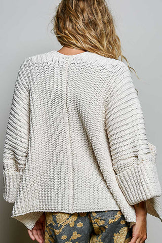 A woman wearing a chunky cream knit cardigan over a cream lace camisole and cream floral pants, photographed against a plain light gray studio background.