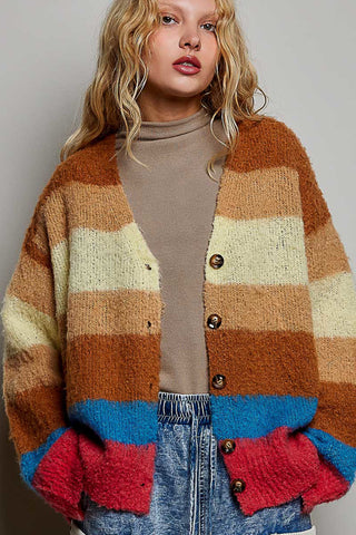 A woman wearing a chunky knit cardigan with wide horizontal stripes in brown, beige, and cream, blue, and red, layered over a beige turtleneck and blue jeans.