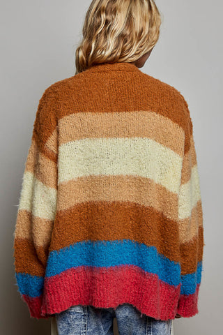 A person viewed from behind wearing a fuzzy knit cardigan with wide horizontal stripes in rust brown, cream, tan, blue, and red, paired with blue jeans against a plain gray studio background.