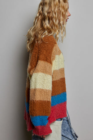 A woman shown from the side wearing a fuzzy striped knit cardigan with horizontal bands of brown, beige, tan, blue, and pink, paired with light-wash denim jeans against a plain gray studio background.
