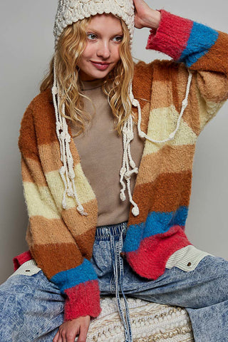 A woman wearing a chunky knit striped cardigan with cream, brown, tan, red, and blue color-blocked stripes, paired with a cream knit beanie and light blue jeans.