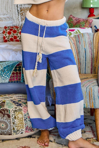 A person wearing loose, ribbed knit pants with wide blue and cream horizontal stripes, standing in a colorful bedroom setting.