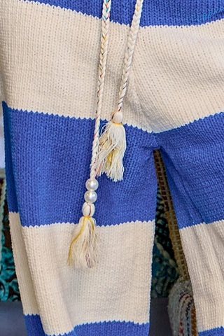 Close-up of a striped knit garment, likely a sweater or cardigan, with wide cream and royal blue horizontal stripes and a cream braided drawstring with tassels and pearl beads.