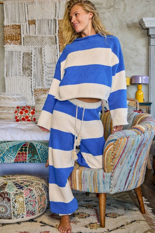 A woman wearing a matching blue and cream striped lounge set, consisting of a cropped long-sleeve top and wide-leg pants with a drawstring, standing in a bohemian-style bedroom next to a colorful striped armchair.