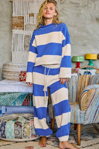 A woman stands barefoot in a bohemian-style room wearing a matching blue and cream striped lounge set consisting of a cropped long-sleeve top and drawstring pants.