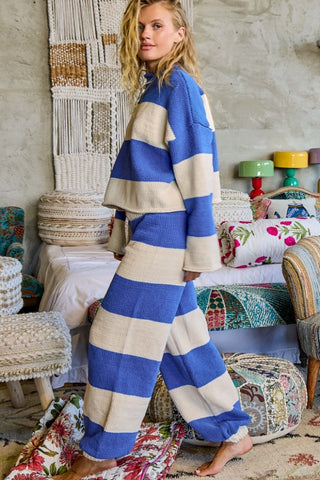 A woman modeling a matching two-piece lounge set with bold horizontal stripes in royal blue and cream, standing in a bohemian-style room filled with woven textiles and patterned cushions.