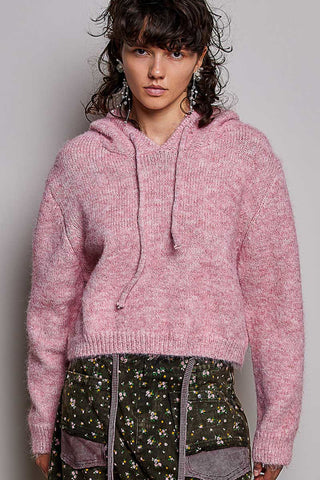 A model wearing a fuzzy, cropped pink knit hoodie with drawstrings, paired with dark green floral patterned pants, against a plain light gray background.