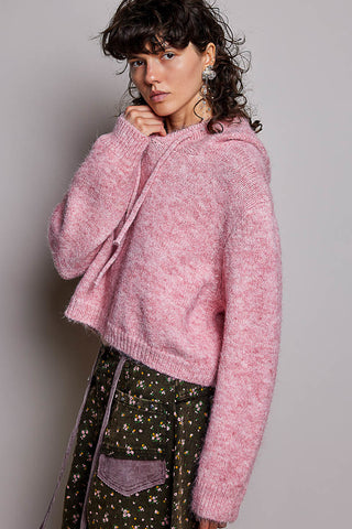 A woman wearing a fuzzy pink cropped knit hoodie sweater and a dark green floral skirt, posing against a neutral gray studio background.