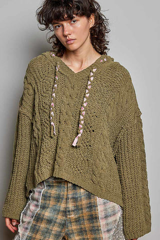 A model wearing a chunky olive green cable-knit cropped hoodie with pink braided drawstrings, paired with plaid pants, against a plain gray studio background.