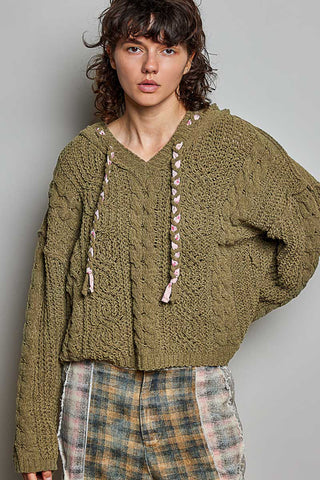 A model wearing a chunky olive green cable-knit cropped hoodie sweater with pink and white tassel drawstrings, paired with a plaid skirt, against a plain gray studio background.