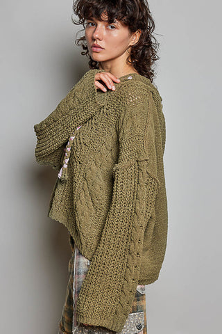 A woman wearing a chunky olive green cable-knit cardigan sweater, shown from a side-back angle against a plain gray studio background.