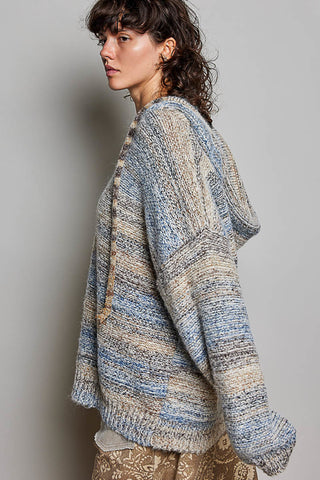 A woman wearing a chunky knit cardigan with horizontal stripes in blue, beige, and cream, shown from a side profile against a neutral gray background.