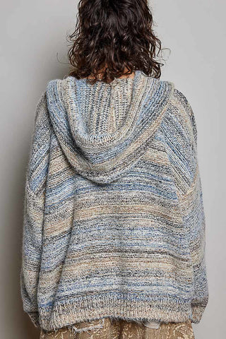 A person shown from the back wearing a chunky knit hooded sweater with horizontal variegated stripes in light blue, beige, and gray, against a plain light gray wall.
