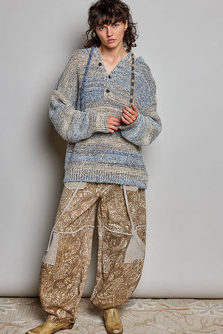 A model stands against a plain light gray wall wearing a chunky knit cardigan in heathered blue and beige tones, paired with loose beige patterned pants and tan boots.