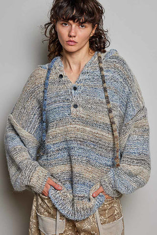 A woman wearing a chunky knit hooded sweater with a blue, beige, and gray marled pattern, paired with patterned pants, against a plain gray background.