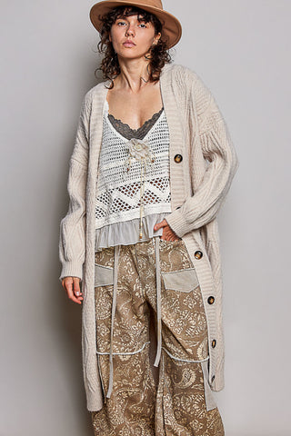 A model wearing a long cream cable-knit cardigan over a white crochet lace-up top, paired with beige paisley trousers and a brown wide-brim hat, against a solid gray studio background.