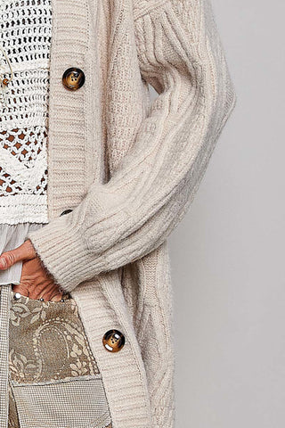 Close-up of a person wearing a chunky cream cable-knit cardigan with brown buttons, layered over a white crochet top and patterned beige trousers, against a plain light gray background.