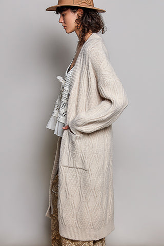 A woman in profile wearing a long cream cable-knit cardigan over a white ruffled blouse, with a brown hat and patterned pants, against a plain light gray studio background.