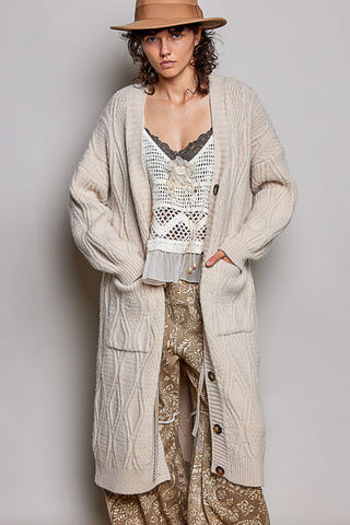 A model wearing a long cream cable-knit cardigan over a white crochet top and patterned pants, styled with a tan hat against a neutral gray background.